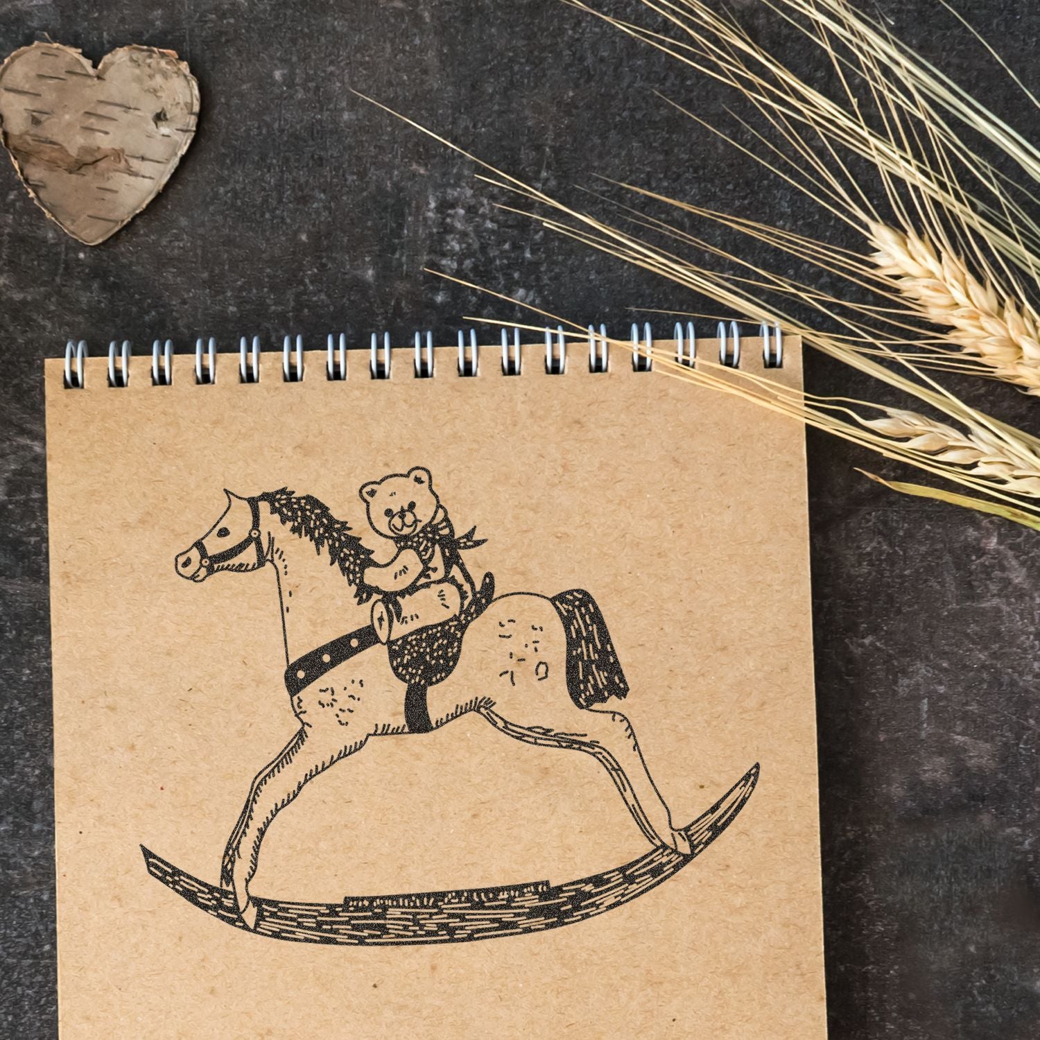 Rustic kraft spiral notebook with teddy bear riding a rocking horse illustration, styled with dried wheat and wood heart on a dark background.