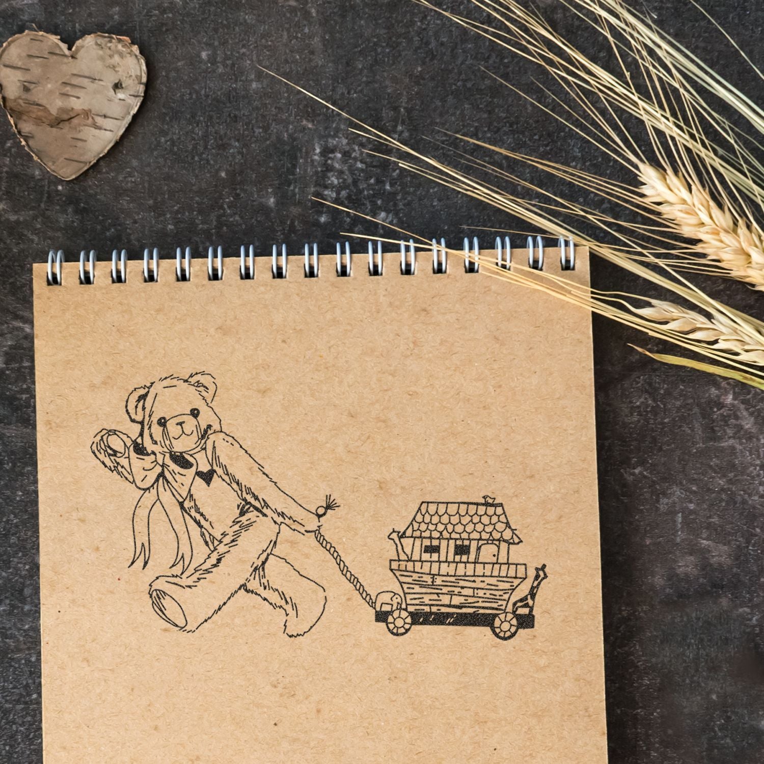 Kraft spiral notebook with cute teddy bear pulling a tiny house cart on a dark rustic background, decorated with dried wheat and a wooden heart.