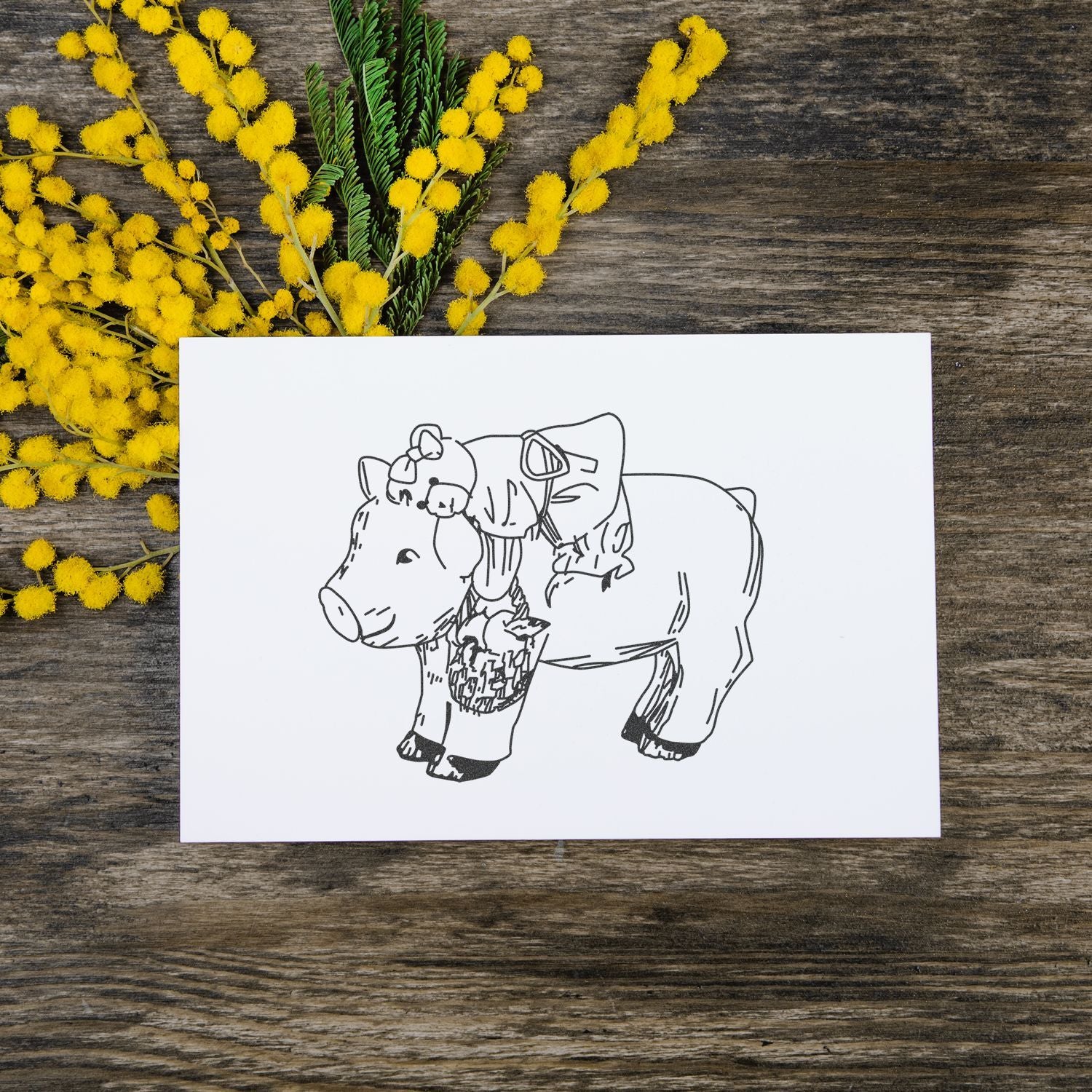 Hand-drawn pig illustration card with yellow mimosa flowers on rustic wooden table.