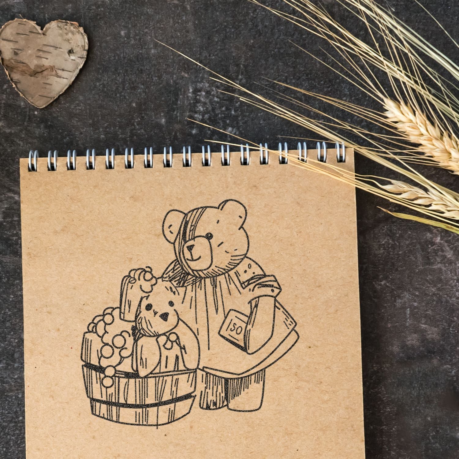 Rustic spiral notebook with cute teddy bear illustration, basket, and wheat on dark textured background.
