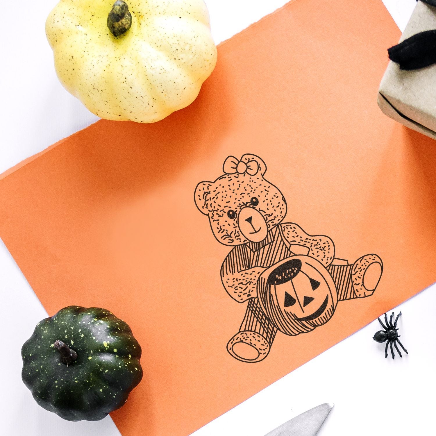 Orange Halloween craft paper with teddy bear pumpkin illustration, surrounded by decorative pumpkins and a spider on a white background