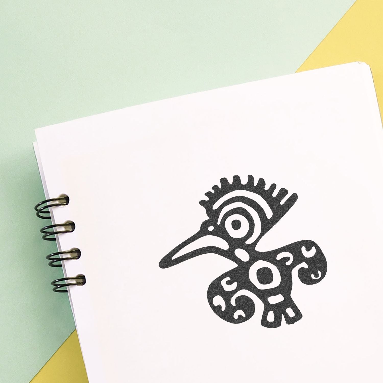 Spiral notebook with a black stylized bird logo on a white page, minimal branding mockup on pastel green and yellow background.
