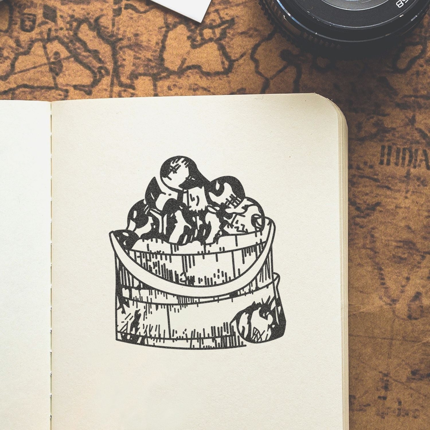 Open sketchbook with black ink dessert illustration on a vintage map background, with camera lens in the corner.