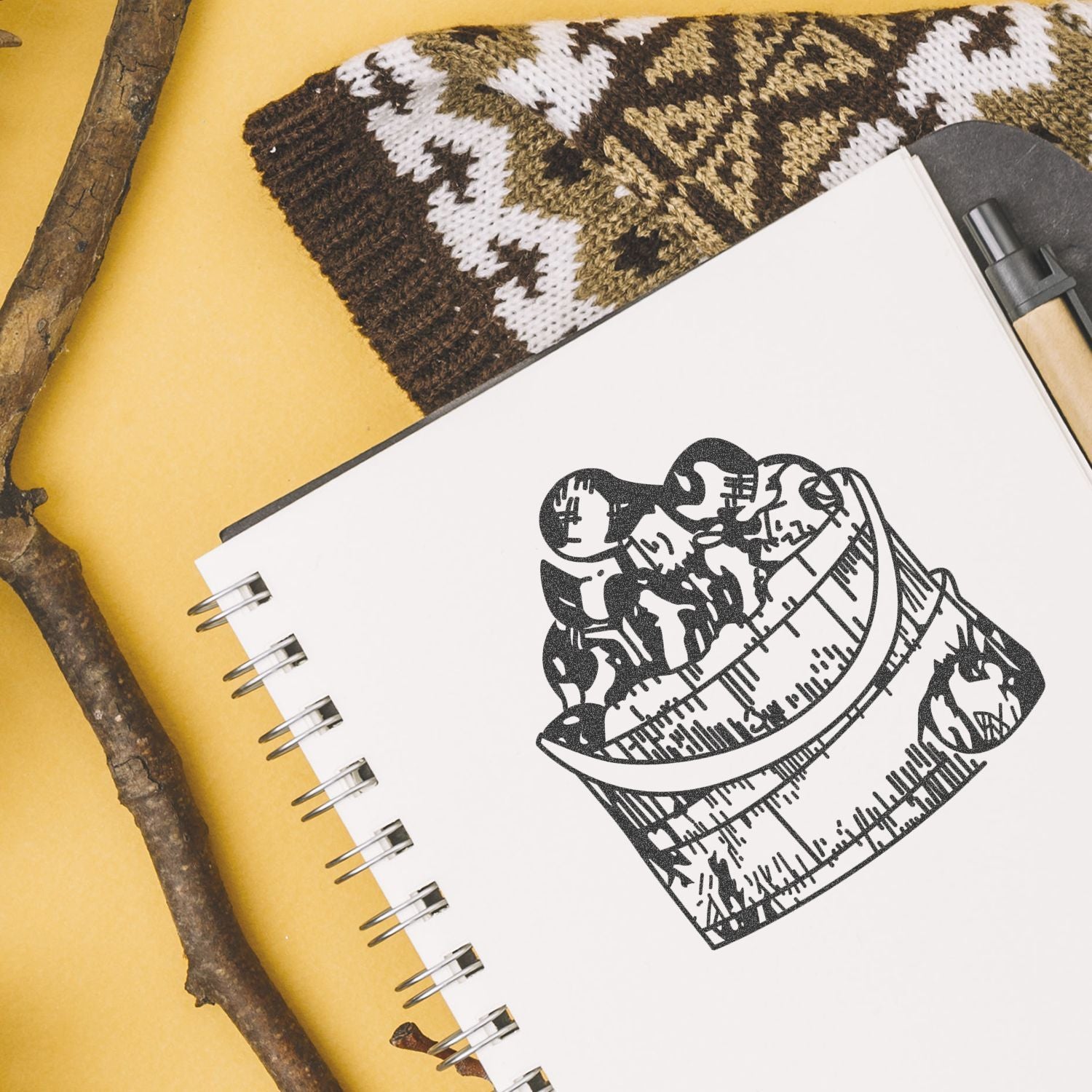 Spiral sketchbook with a black coffee mug illustration on a yellow background, styled with rustic twigs and knitted fabric.