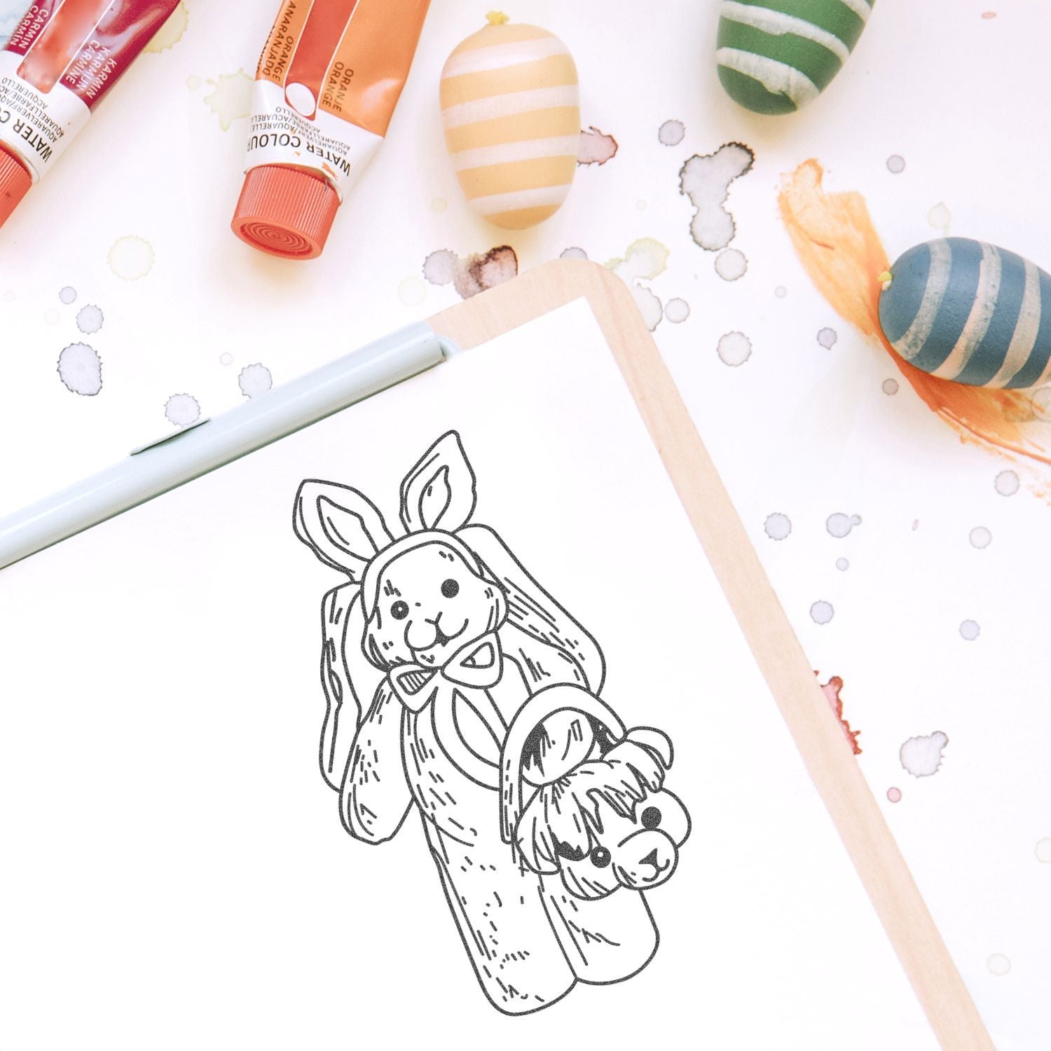 Cute Easter bunny coloring page with watercolor paints, painted eggs, and art supplies on a sketchbook.