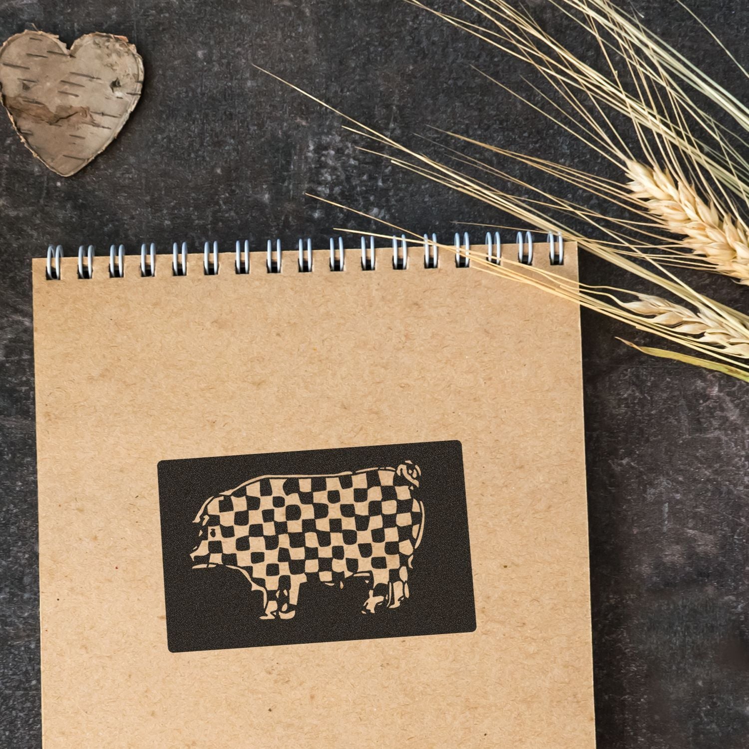 Rustic notebook with a checkered cow silhouette, dried wheat, and a heart-shaped wood slice on a dark background.