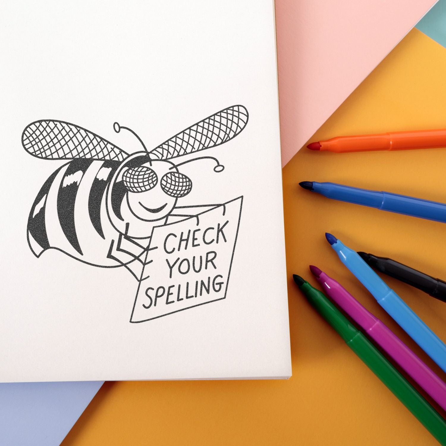 Open notebook with a bee illustration holding a Check Your Spelling sign, surrounded by colorful markers on a pastel background.