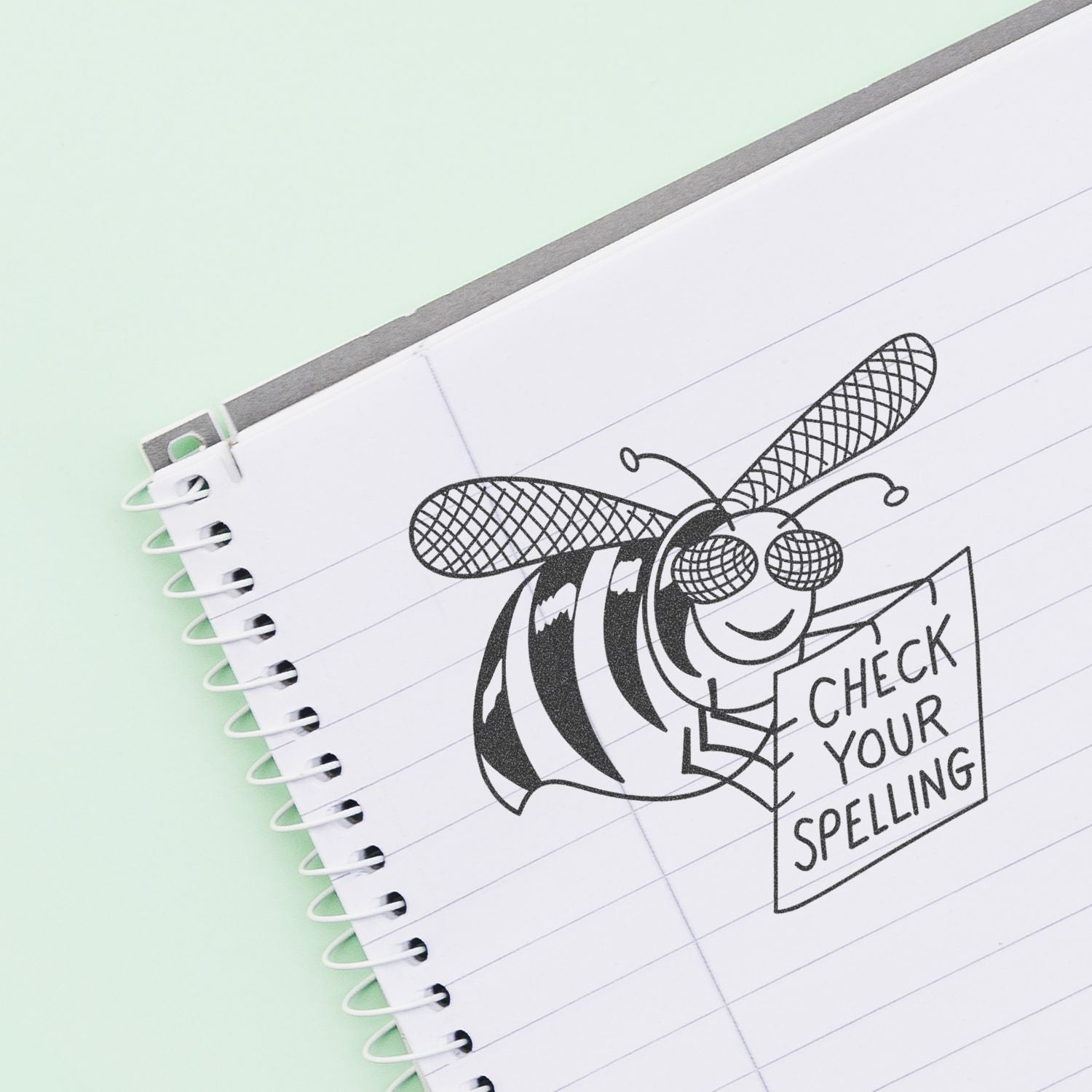 Spiral notebook with a cute bee illustration holding a Check Your Spelling sign on lined paper.