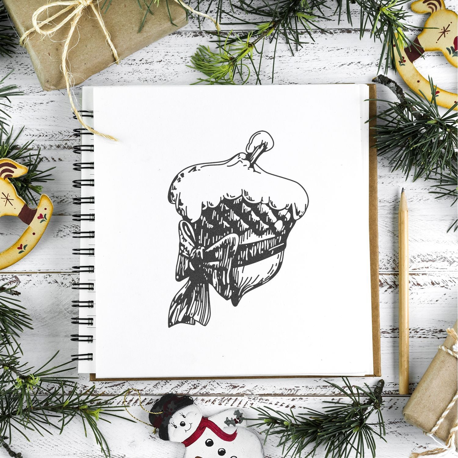 Top view of a sketchbook with black and white Christmas pudding illustration surrounded by festive holiday decorations and pine branches.