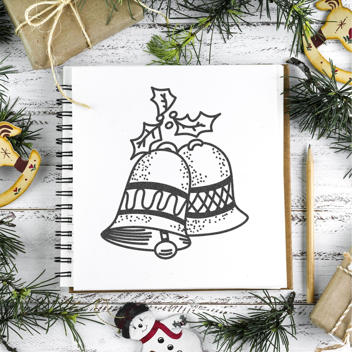 Christmas bells illustration in a sketchbook with festive pine branches and holiday gift wrapping on a rustic white wooden background.