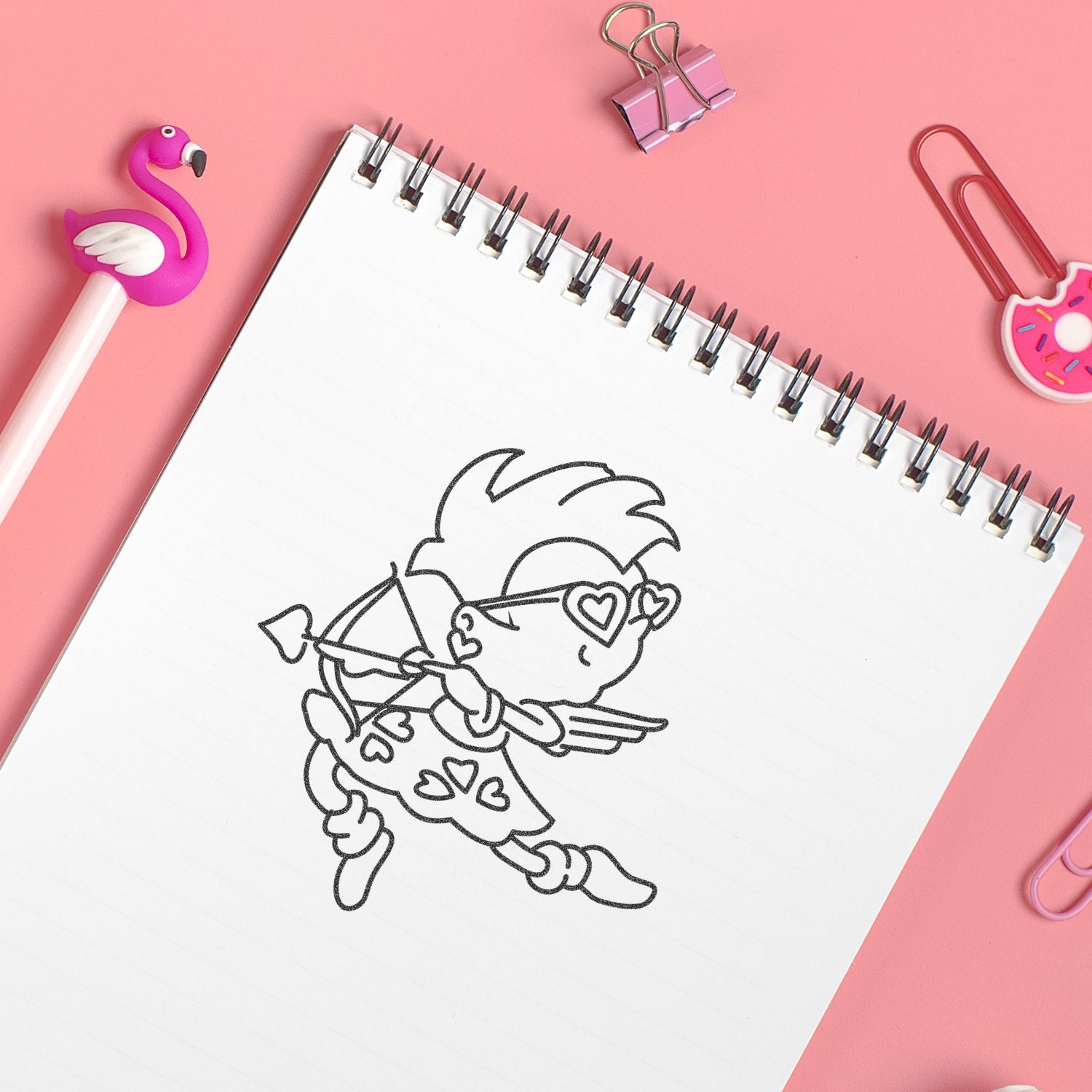 Cute cupid sketch in a spiral notebook on a pink background with colorful stationery accessories and paper clips.