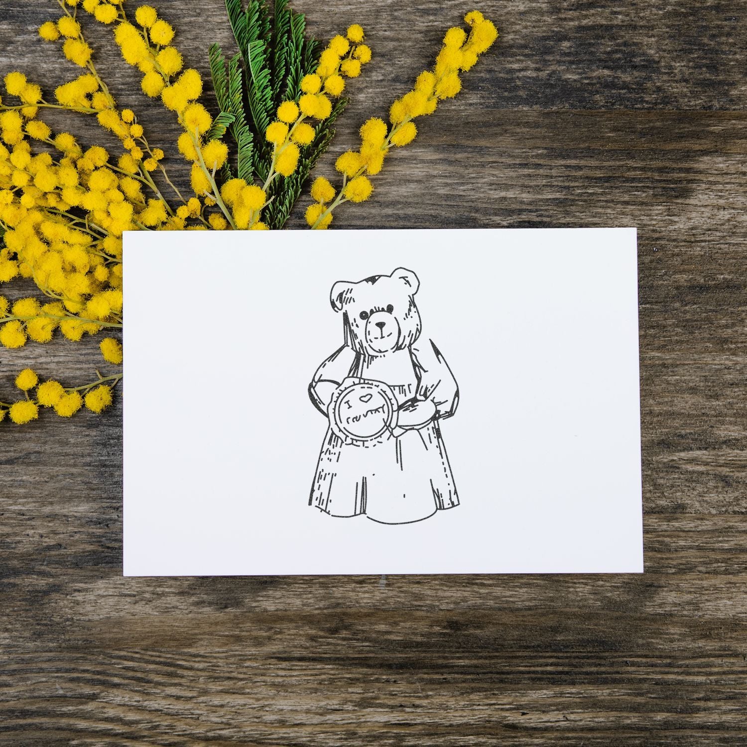 Teddy bear illustration on a white greeting card with yellow mimosa flowers on a rustic wooden table.