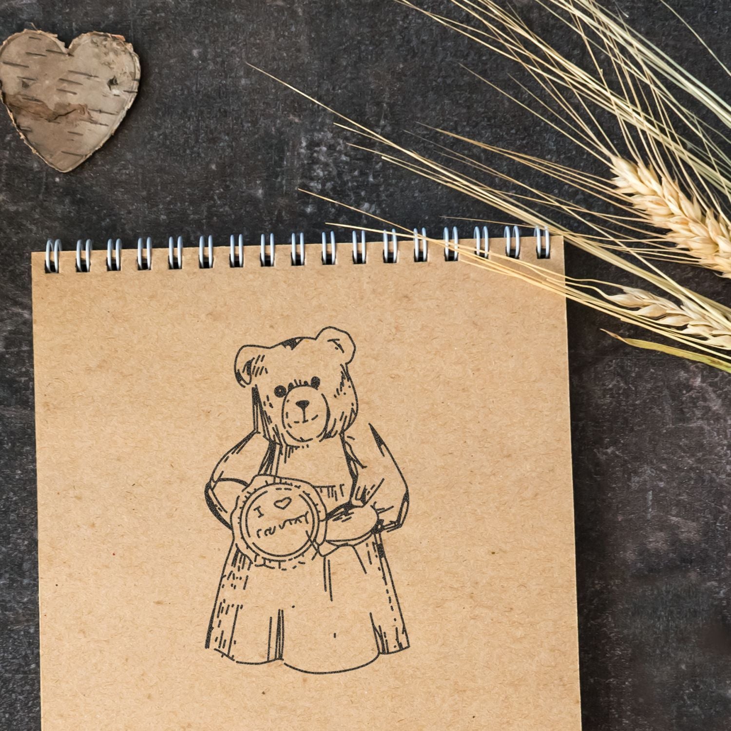 Rustic kraft paper spiral notebook with cute bear illustration, dried wheat, and birch heart on a dark background