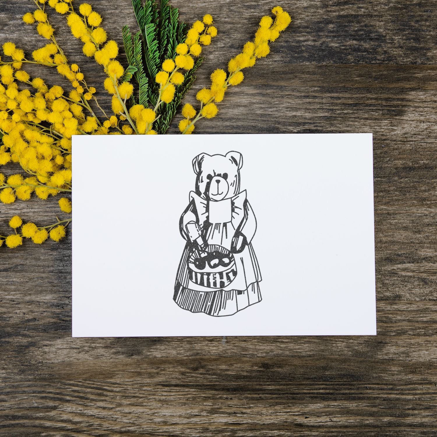 Hand-drawn teddy bear greeting card on rustic wood with yellow mimosa flowers.