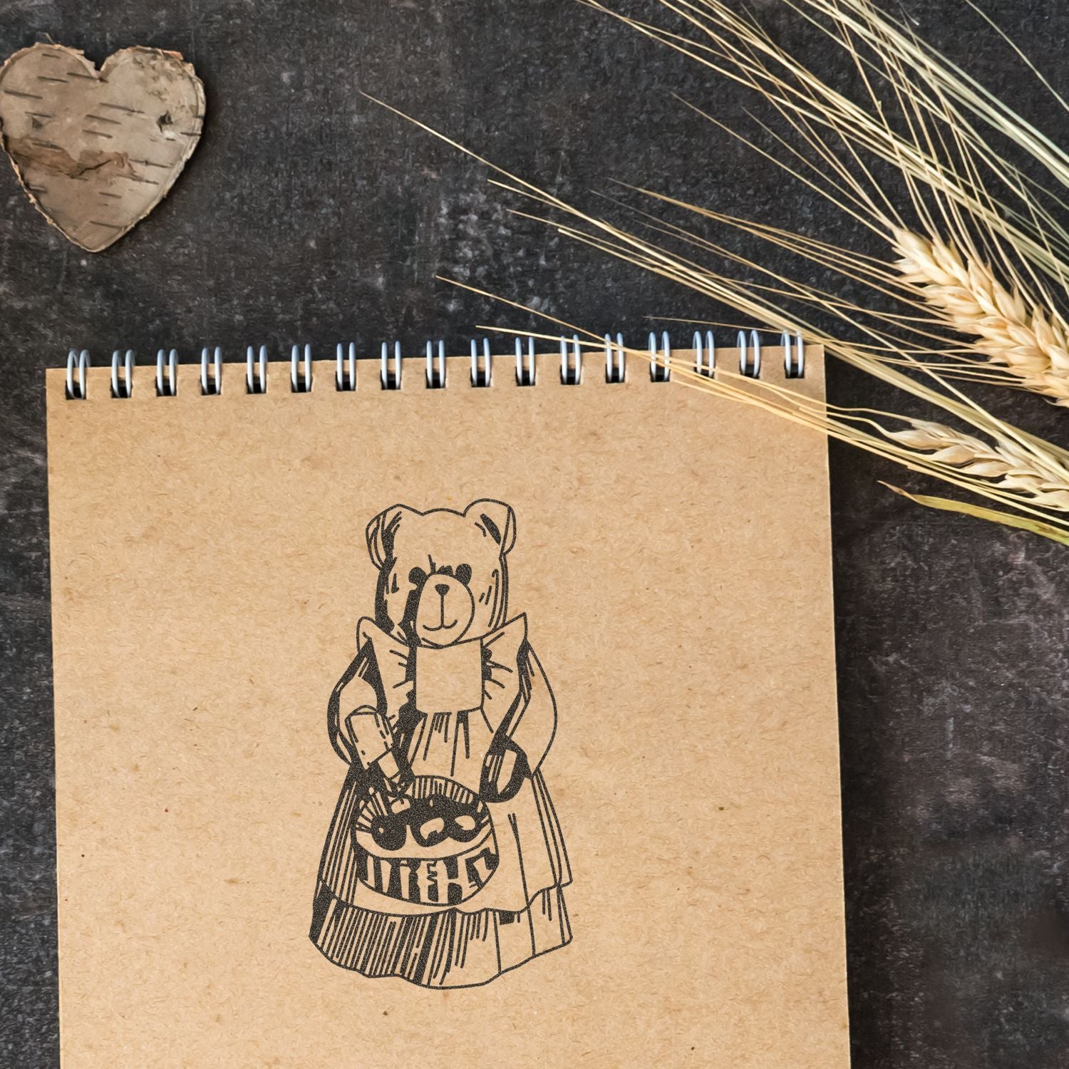 Spiral kraft paper notebook with teddy bear illustration, rustic decor, and dried wheat stalks on a dark background