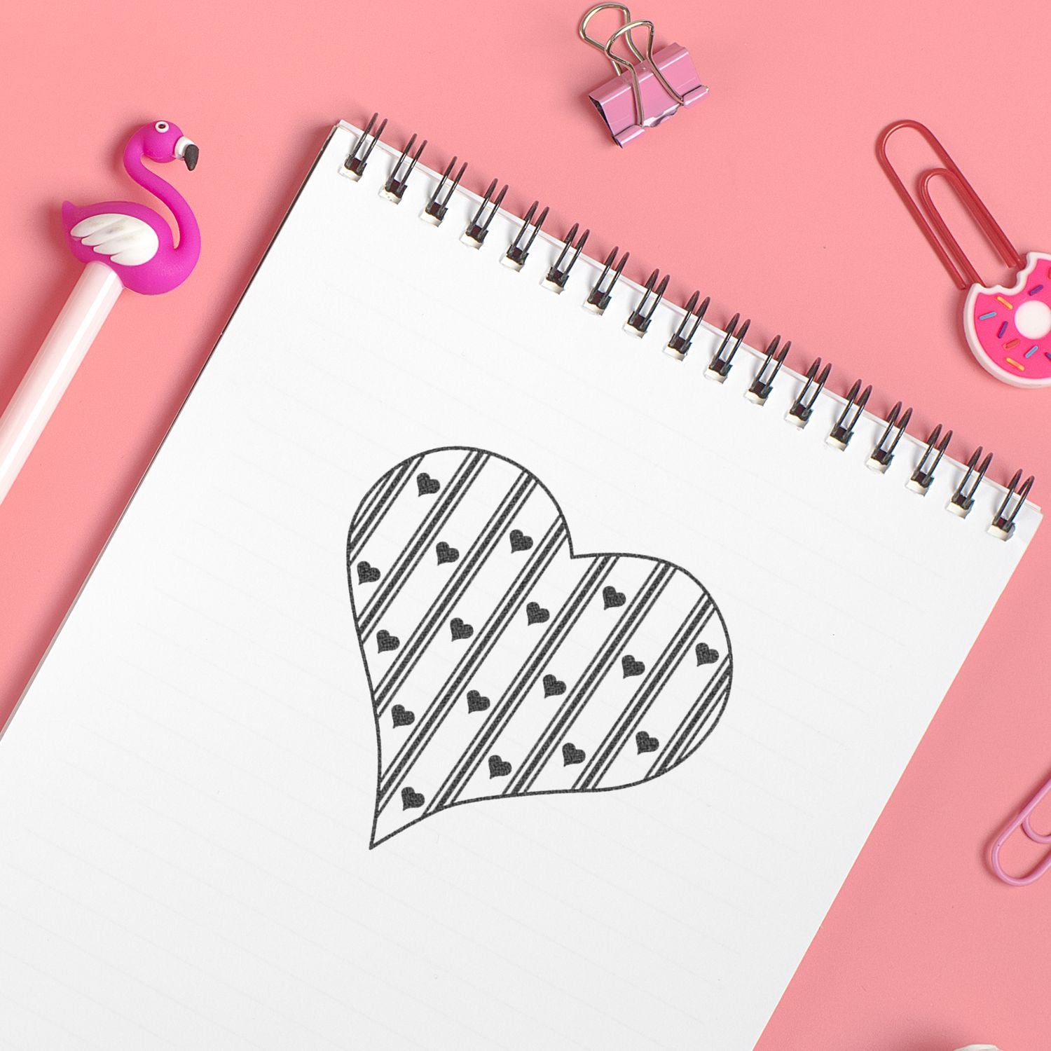 Pink stationery flat lay with spiral notebook showing a heart doodle, flamingo pen topper, and cute binder clips on a pastel pink background.