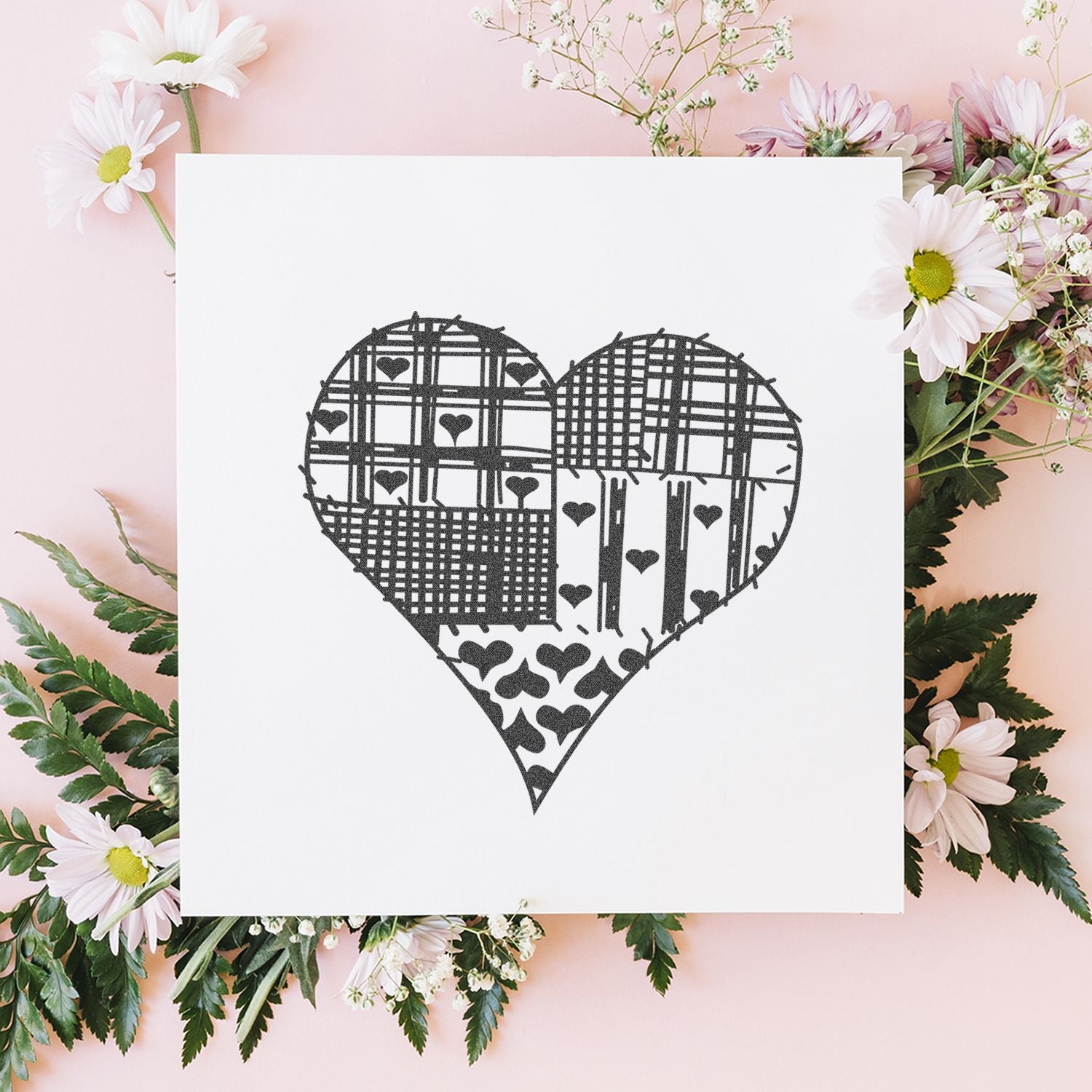 White card with black patchwork heart design surrounded by pastel pink daisies and greenery on a soft pink floral background.