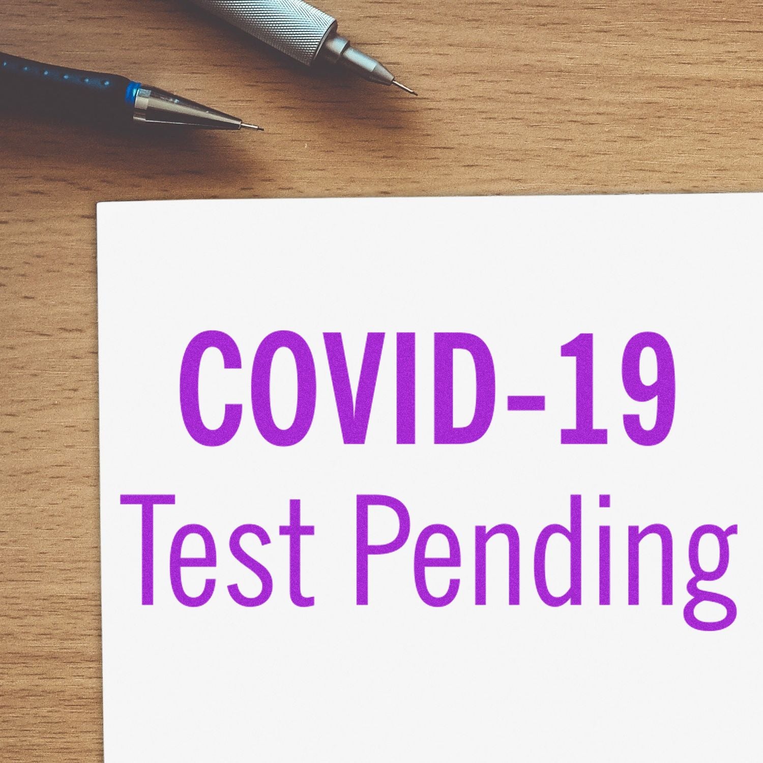 COVID-19 test pending message on white card over wooden desk with pens, indicating awaiting test results.