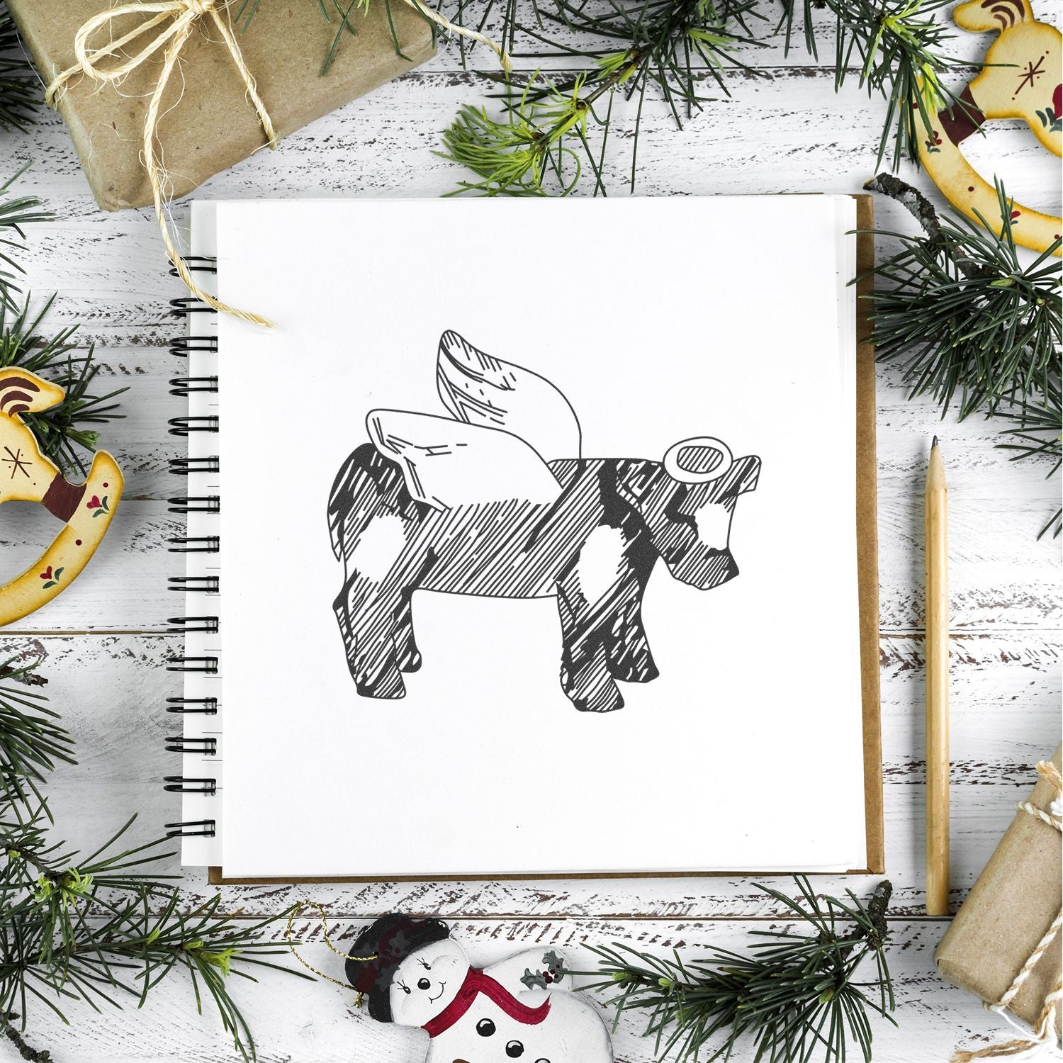 Holiday flat lay with sketchbook featuring a winged cow illustration, pine branches, wrapped gifts, and festive Christmas ornaments.