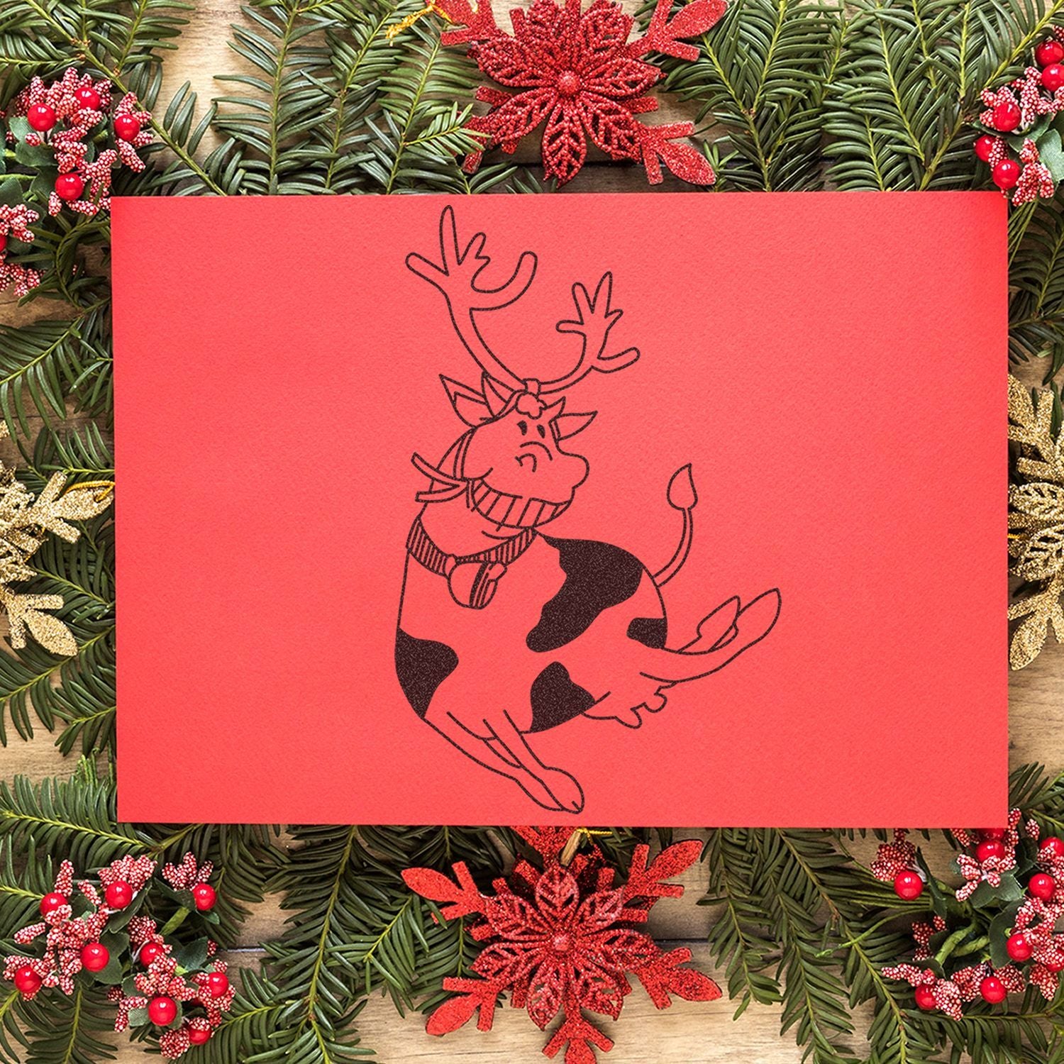 Festive Christmas greeting card with a cute cow wearing reindeer antlers, surrounded by pine branches, red ornaments, and holiday decorations.
