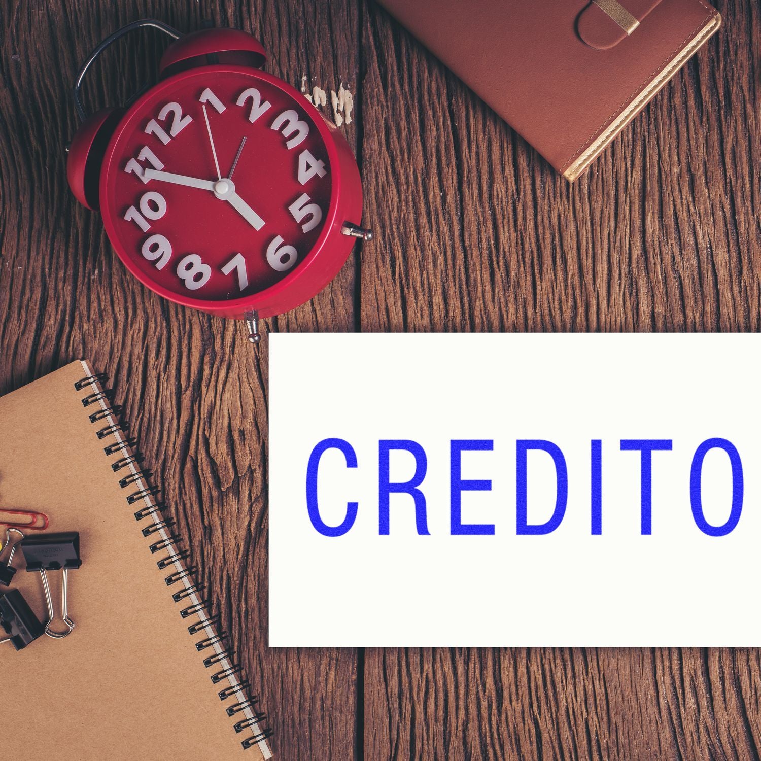 Red alarm clock and notebook on wooden desk with CREDITO text, symbolizing payment deadline and financial credit reminders today.