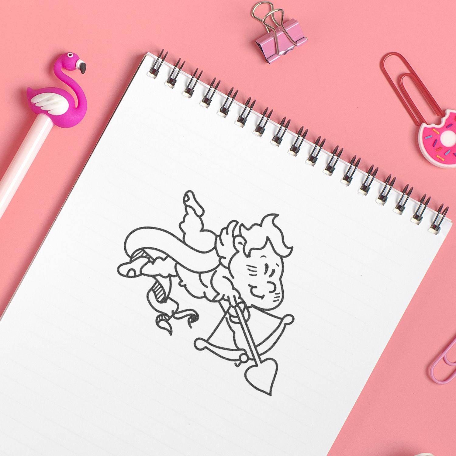 Pink stationery flat lay with spiral notebook featuring a cute Cupid drawing, flamingo pen, and colorful paper clips on a pastel pink background.