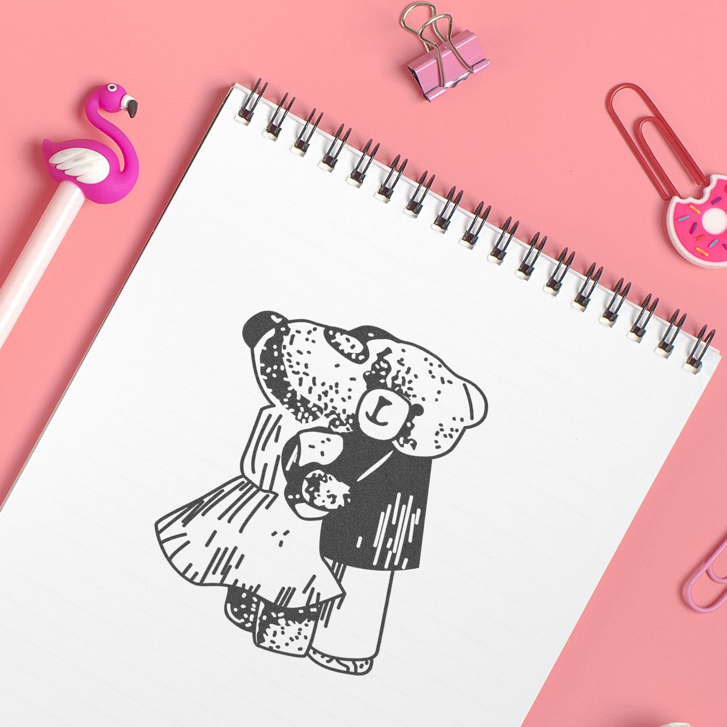 Pink desk flat lay with spiral notebook featuring a cute teddy bear illustration, flamingo pen, donut paper clips, and pastel stationery accessories.