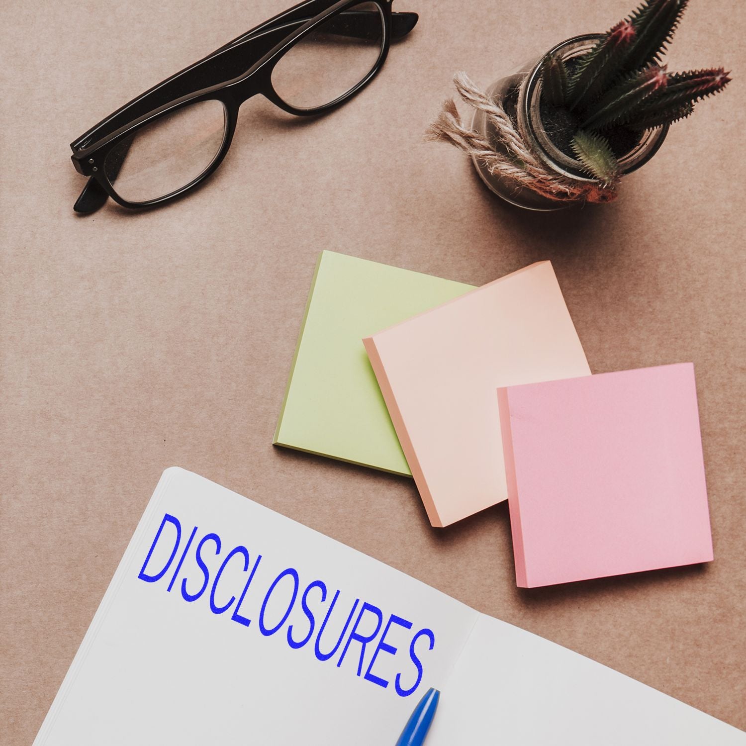 Desk flat lay with eyeglasses, sticky notes, plant, and notebook labeled Disclosures for office or content compliance planning