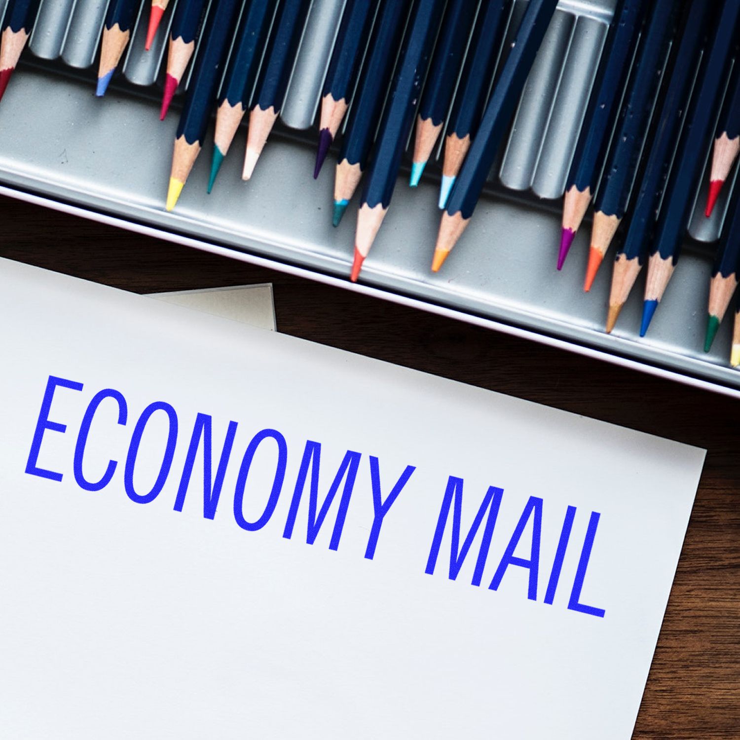 Economy mail concept with ECONOMY MAIL on white paper, colored pencils in tray on wooden desk for postal shipping.