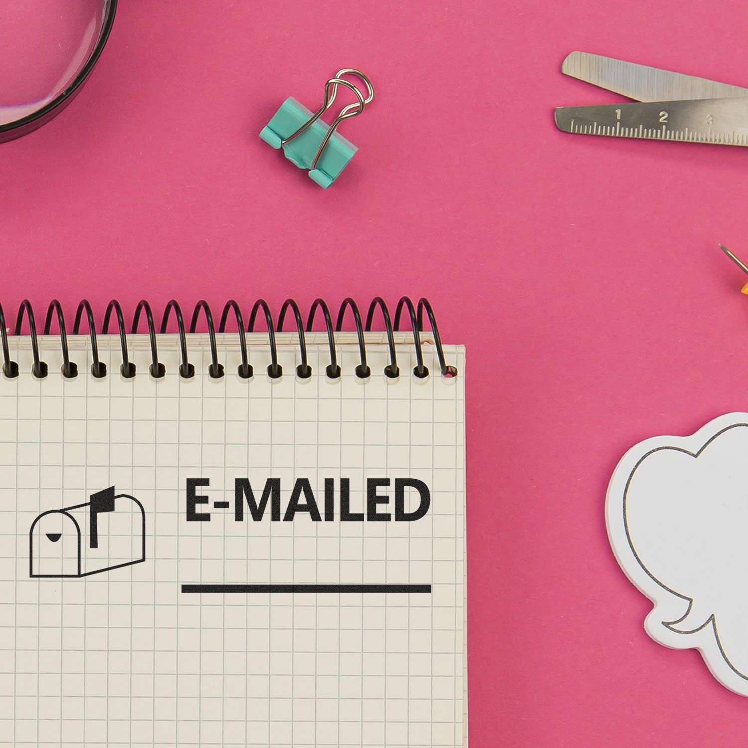 Notebook with E-MAILED text on a pink desk surrounded by stationery supplies and office tools