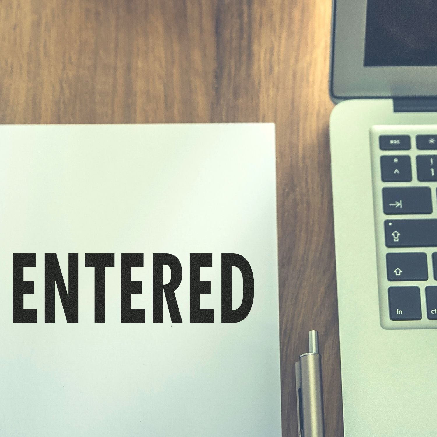 Word ENTERED on white paper next to a laptop keyboard and pen on a wooden desk.