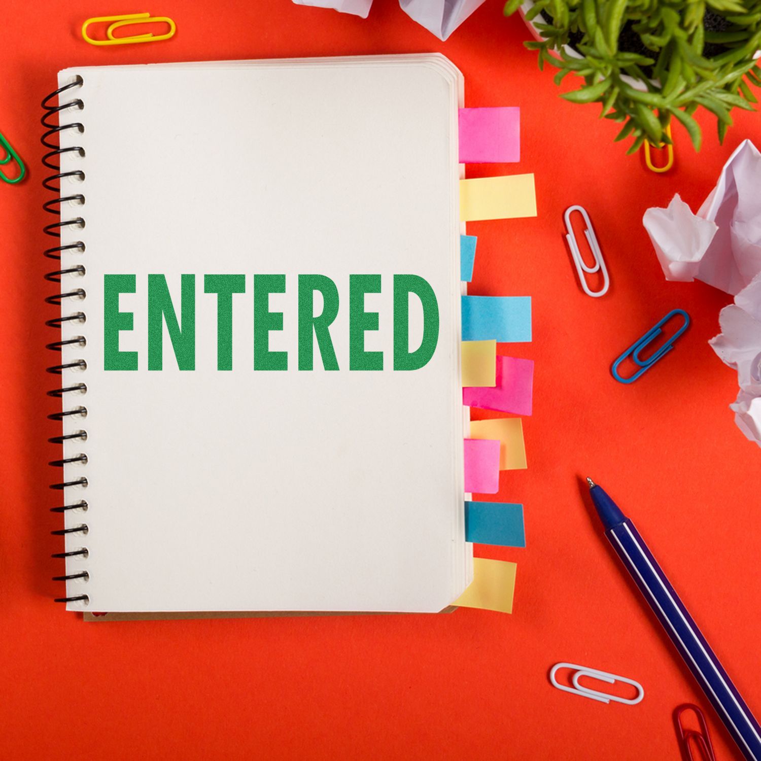 Notebook labeled ENTERED with paper clips, sticky notes, and pen on red deskdocument entry concept