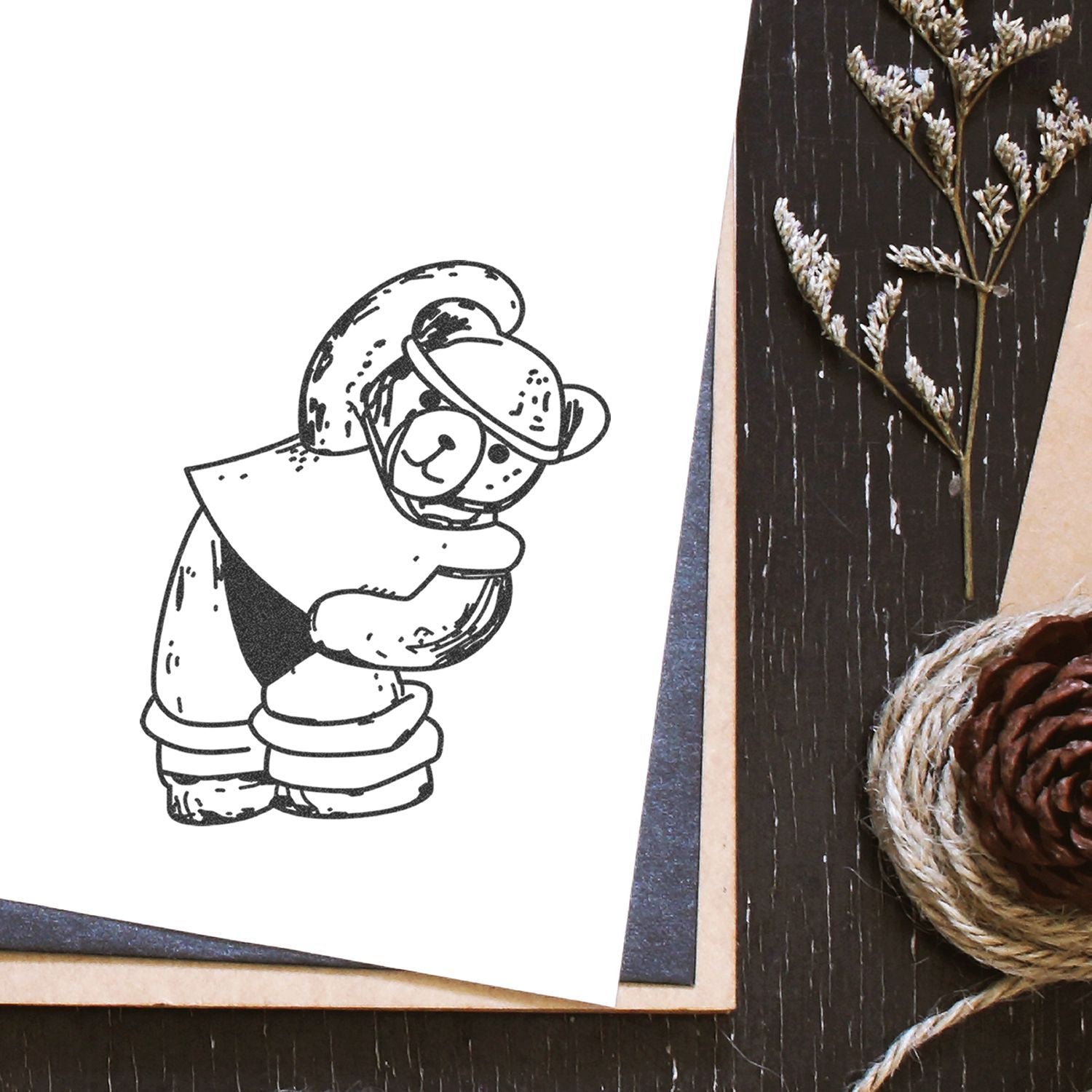 Cute hand-drawn teddy bear illustration on a handmade greeting card with rustic pinecone, twine, and dried floral decor on a dark wooden table.