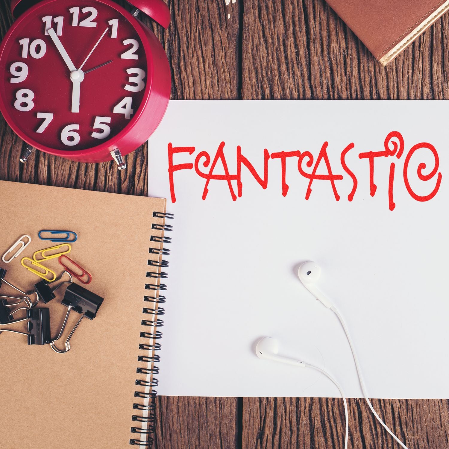 Red alarm clock, notebook with fantastic text, earbuds, and office stationery on a wooden desk.