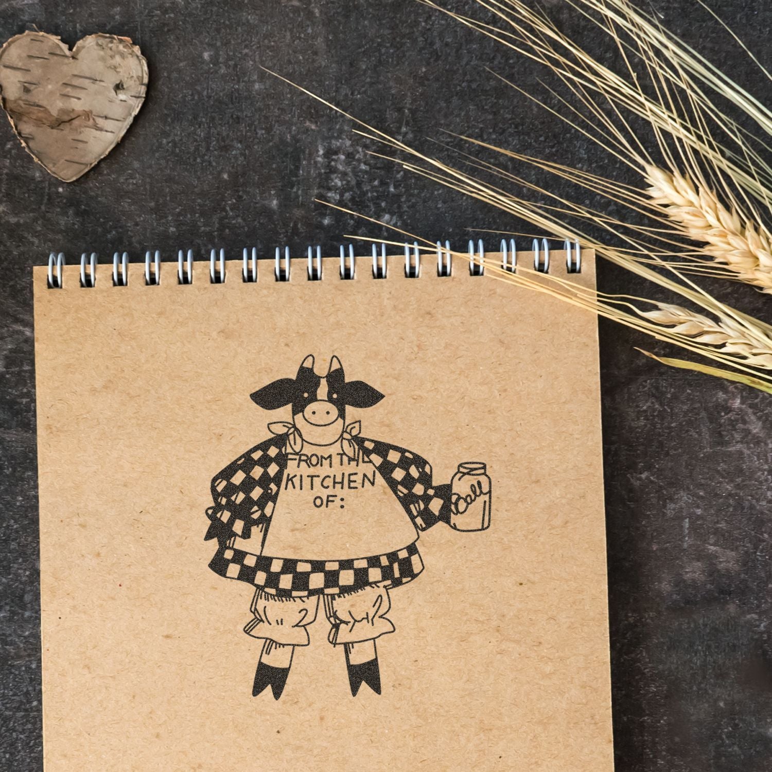 Rustic kitchen notebook with cow illustration, dried wheat, and birch heart on dark background.