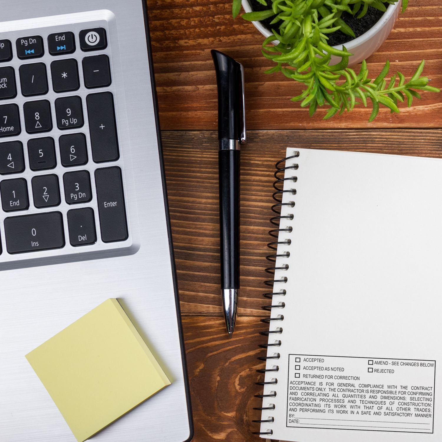 Top view of laptop keyboard, notebook, pen, sticky note, and potted plant on a wooden office desk workspace