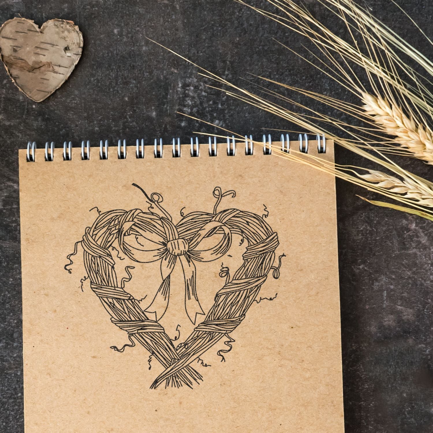 Rustic heart-shaped twine wreath drawing on a kraft paper spiral notebook, styled with wheat stalks and a birch heart on a dark background.