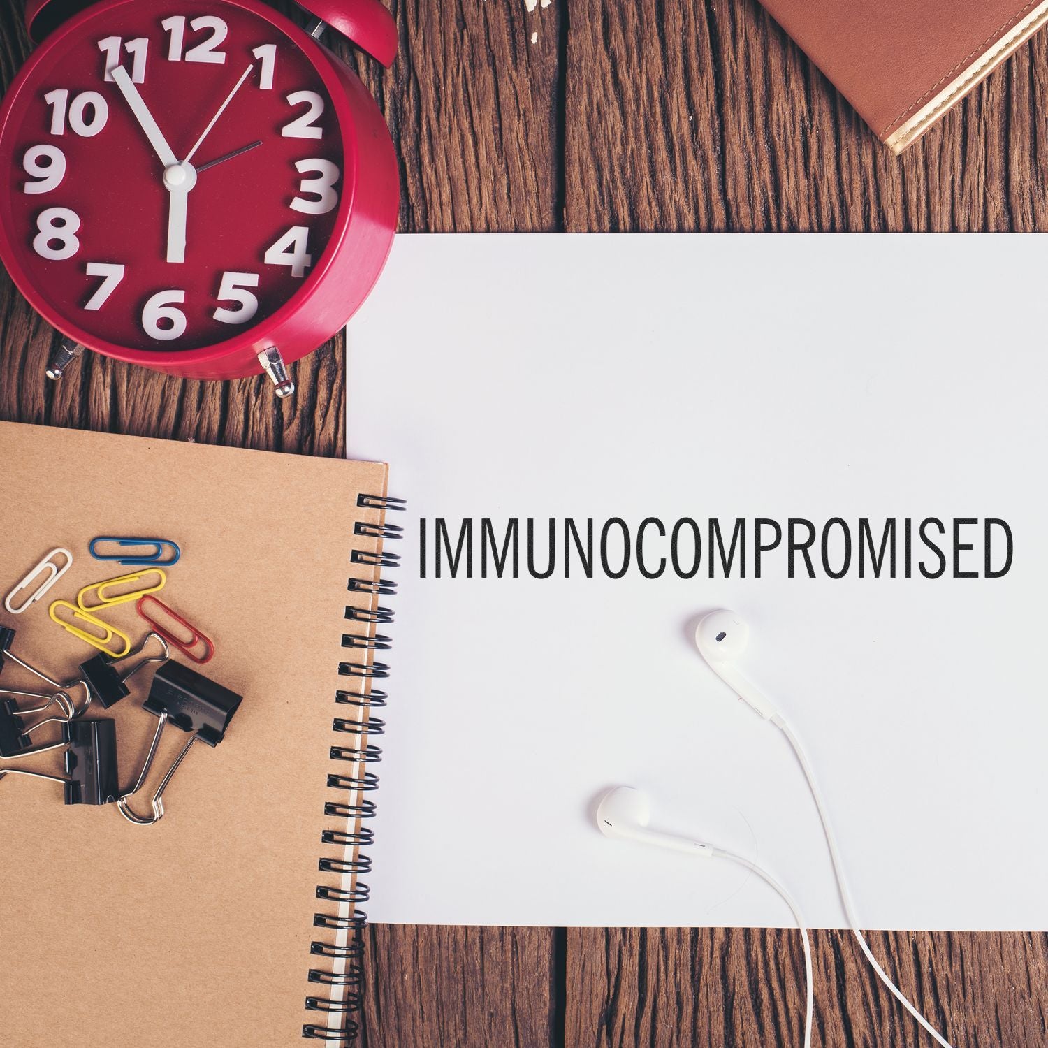 Desk flat lay with red alarm clock, notebook, earbuds, and IMMUNOCOMPROMISED text on white paper, concept for health awareness and time management.