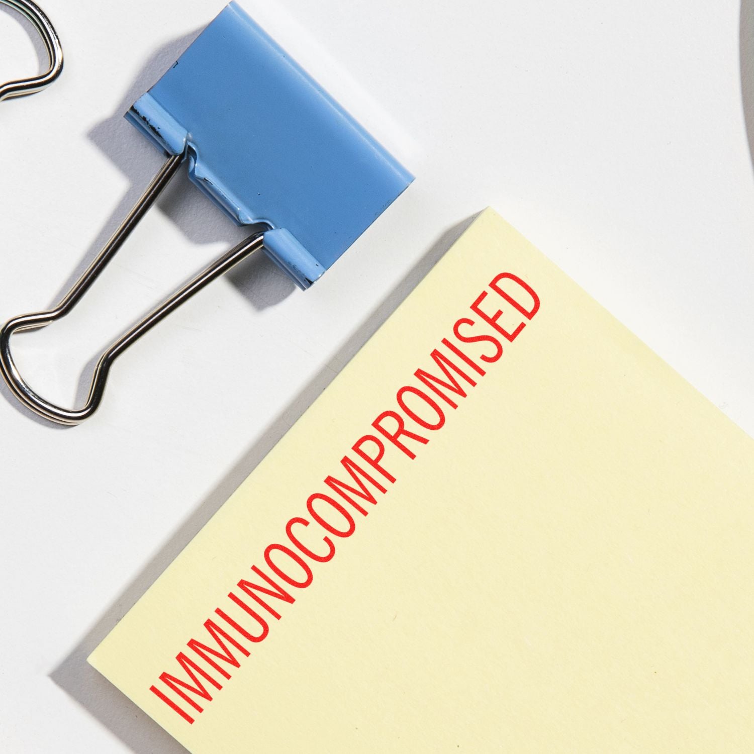 Blue binder clip holding a note labeled immunocompromised on a white background, concept of immune system health and medical vulnerability.
