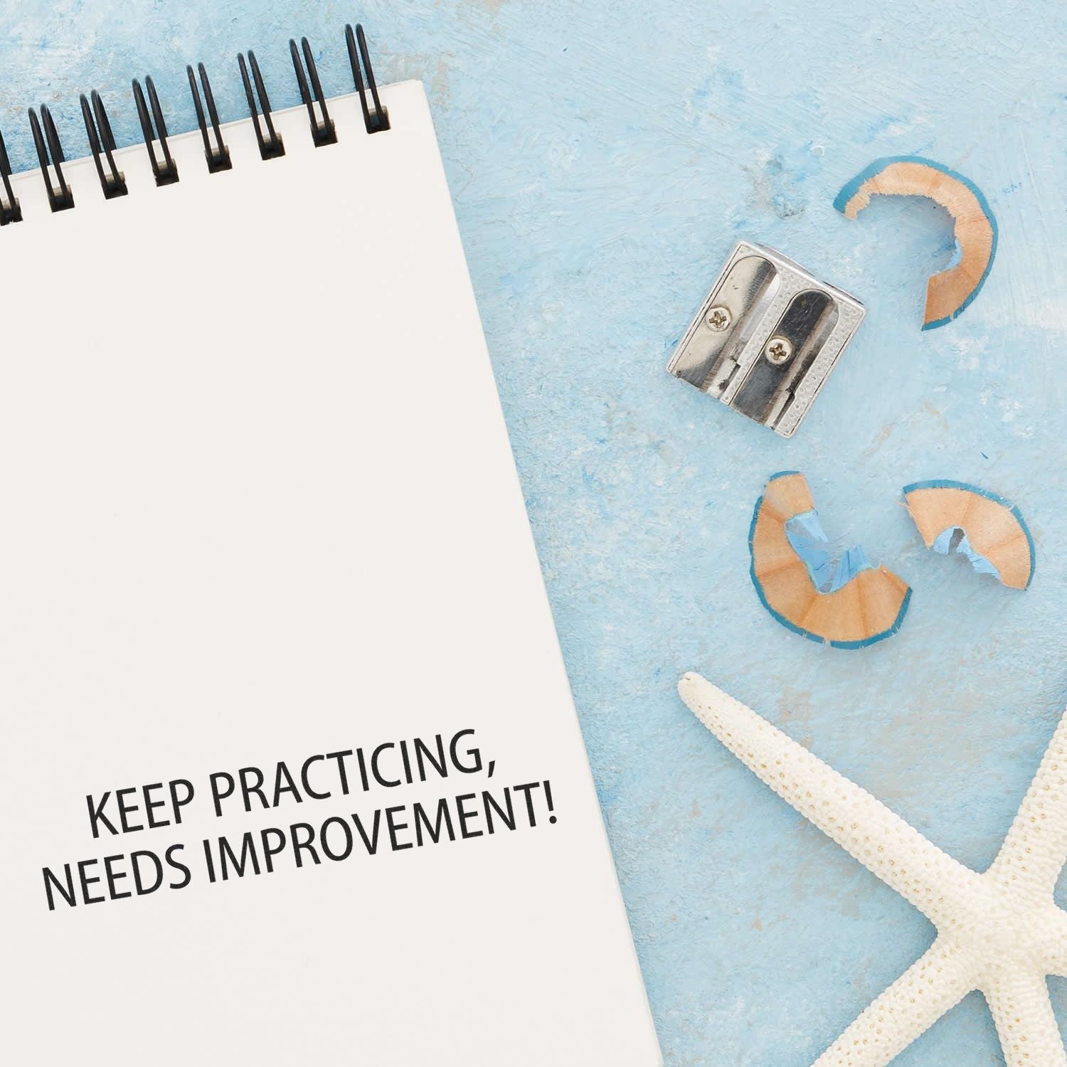 Notebook with Keep Practicing, Needs Improvement message on blue background with pencil sharpener and starfish, inspirational learning concept.