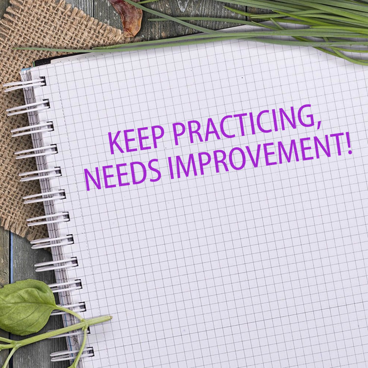 Notebook with purple Keep Practicing, Needs Improvement! text on graph paper, surrounded by greenery and rustic desk decor.