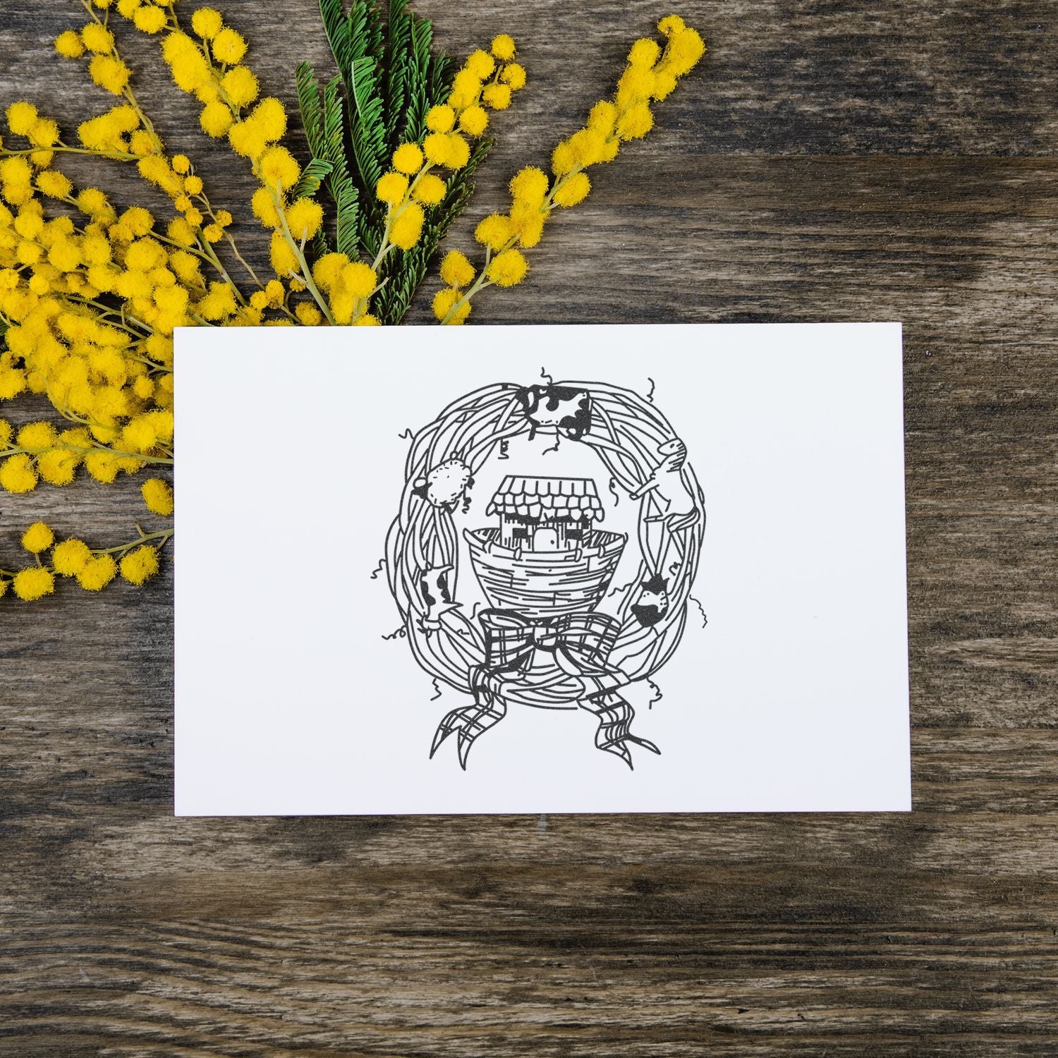 White greeting card with nautical wreath illustration on rustic wooden background, decorated with yellow mimosa flowers.