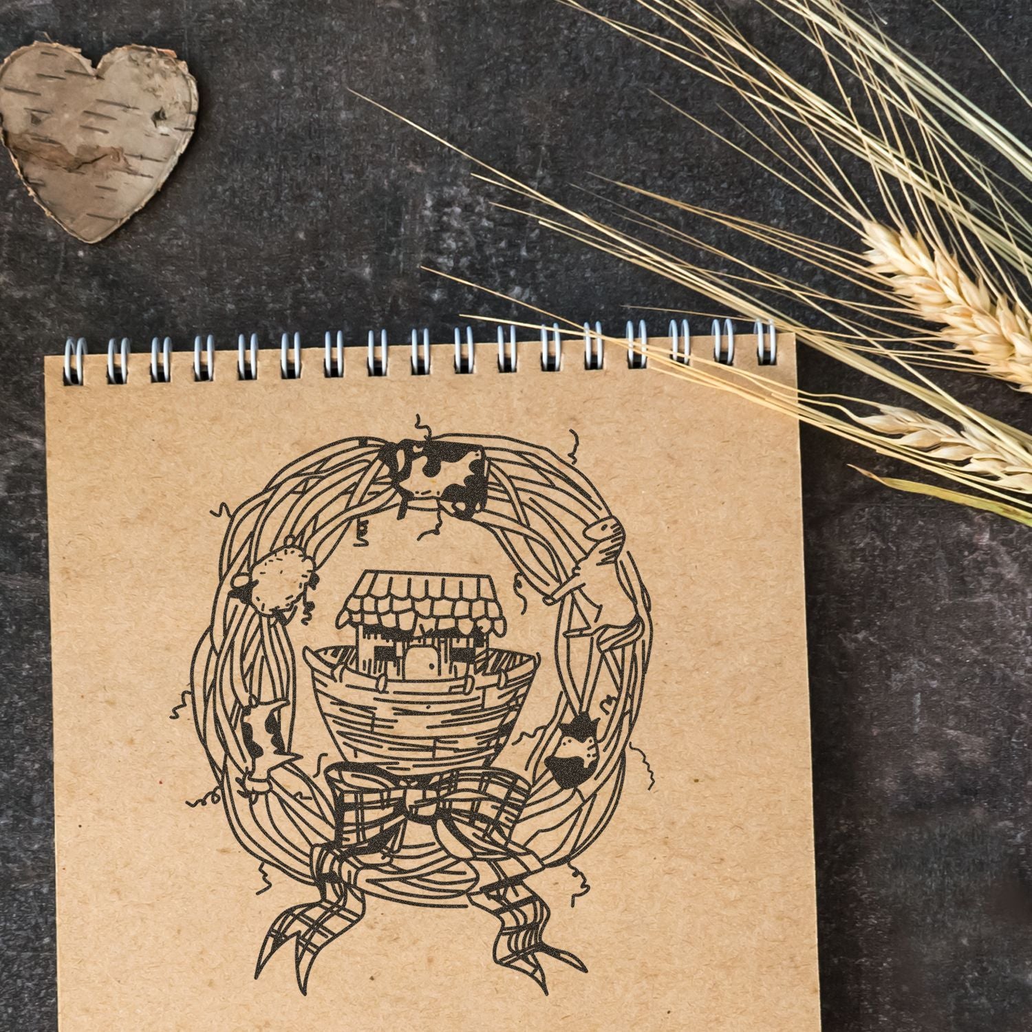 Rustic spiral notebook with farmhouse birdhouse illustration, wheat stalks, and wooden heart decor on a dark background.
