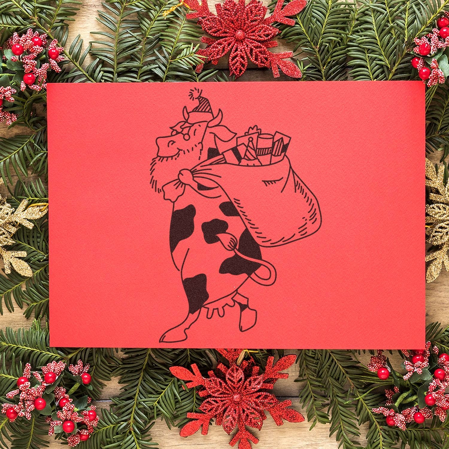 Festive Christmas card with a whimsical cow carrying gifts on a red background, surrounded by holiday pine branches and red snowflake decorations.