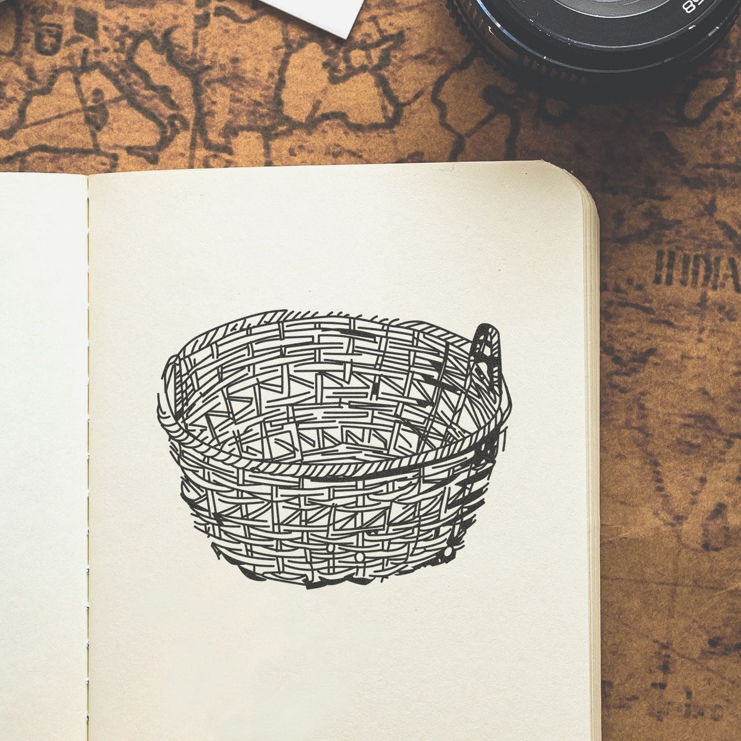 Open sketchbook with a black line drawing of a woven basket on a cream page, styled on a vintage map background with a camera lens in the corner.