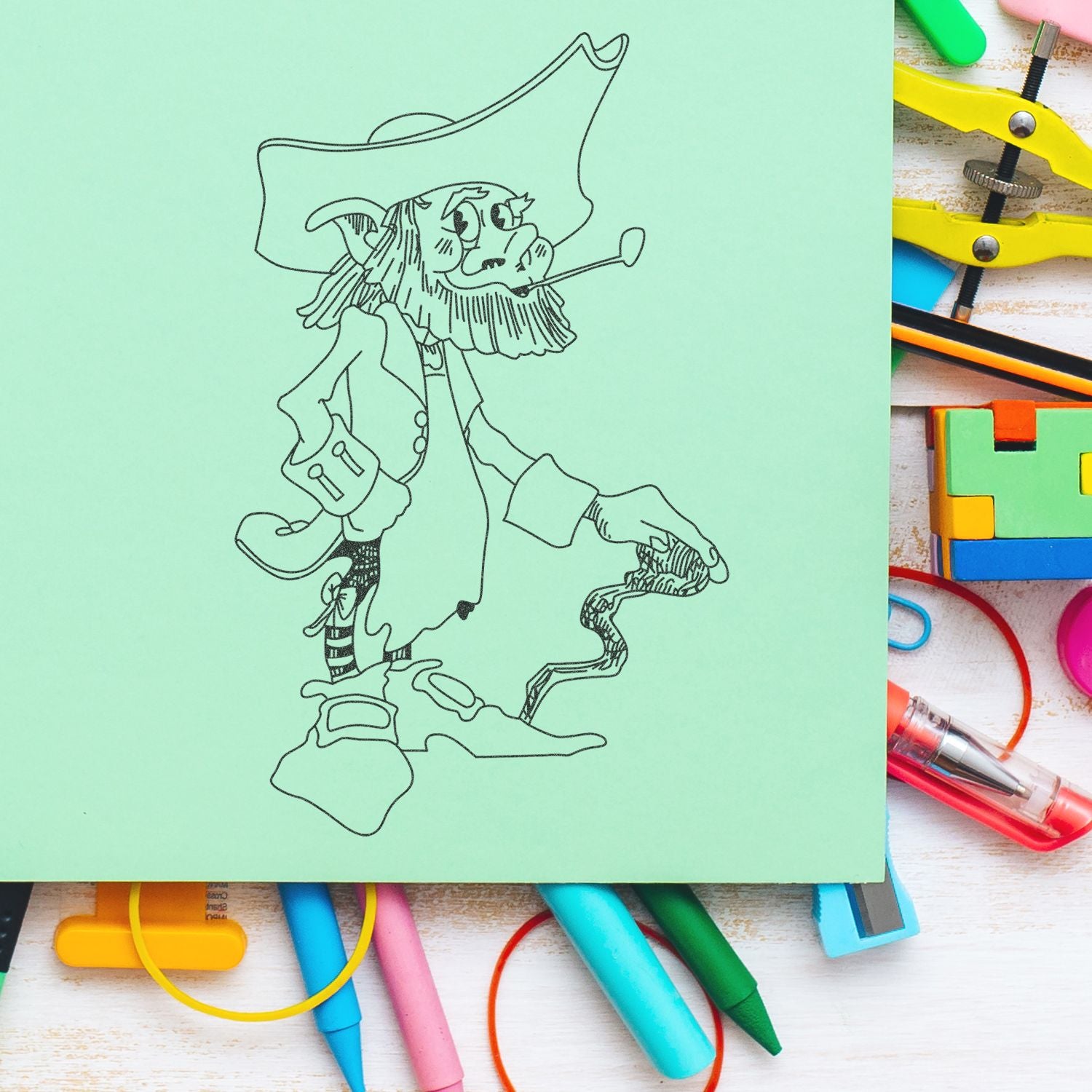 Pirate coloring page on mint green paper surrounded by colorful school supplies and art tools on a white desk.