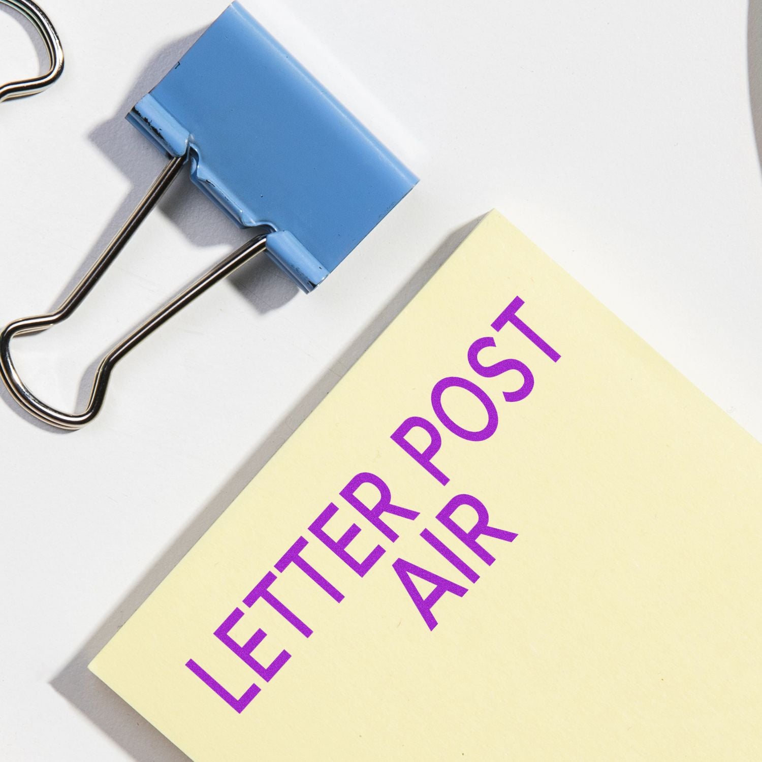Close-up of a binder clip holding a sticky note labeled Letter Post Air on a white background.