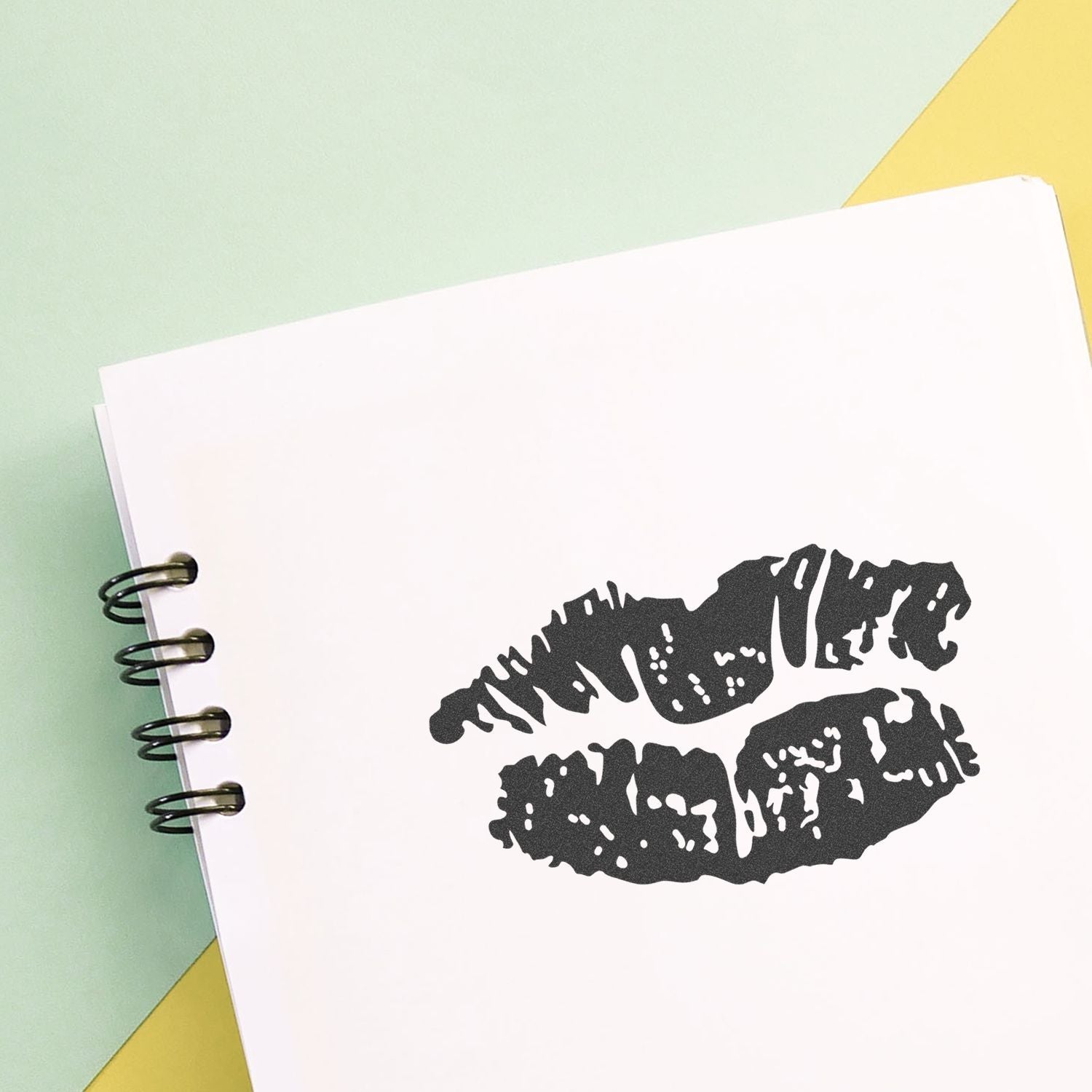 Spiral notebook with black lipstick kiss print on a pastel green and yellow background.