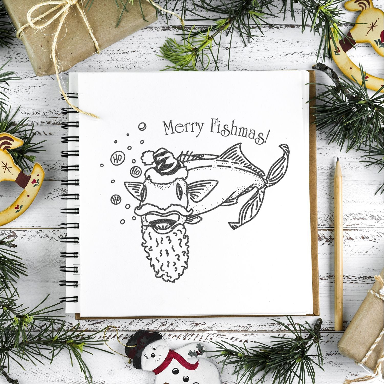 Holiday sketchbook with Merry Fishmas Christmas fish drawing, surrounded by pine branches, wrapped gifts, and festive decorations on a rustic white table.
