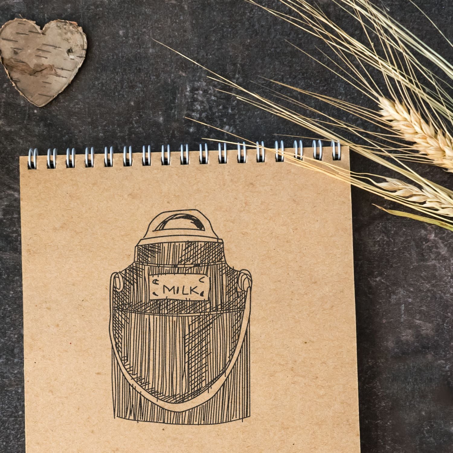 Rustic kraft notebook with a hand-drawn milk can illustration, wheat stalks, and a wooden heart on a dark textured background.